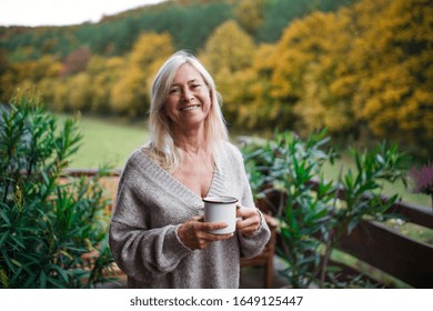 Senior woman with coffee standing outdoors on terrace, looking at camera.