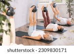 Senior woman in company of elderly friends and daughter perform Pilates exercises with softball. Visitior yoga class lying on back, raised legs perpendicular to floor and clamped softball in feet