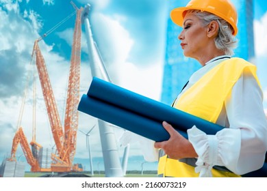 Senior Woman Construction Manager With Blue Print At Wind Propeller Turbines Site. Older Female Architect With Plan For Renewable Sustainable Engineering. Copy Space