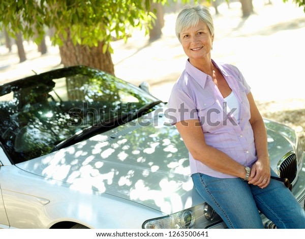 Senior woman with\
convertible car on road trip drive in countryside sitting on bonnet\
smiling at camera