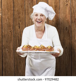 Senior Woman Cook Holding A Tray With Muffins Against A Wooden Background