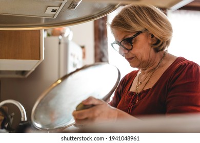 Senior Woman Cooking Lunch In The Kitchen - Mature Woman In The Kitchen Lifting The Lid Of The Pot To Check The Cooking -  Boomer Woman Busy Cooking Lunch For The Family