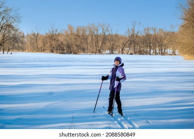 Senior Woman Cross-country Skier Smiling At Camera In Late Afternoon In Midwest; Fresh Snow In Front Of Her; Bare Trees And Blue Sky In Background