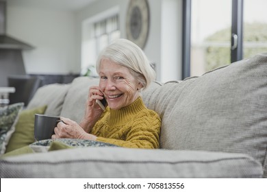 Senior Woman Is Curled Up On The Sofa In Her Home With A Cup Of Tea While She Enjoys A Phonecall On A Smartphone.