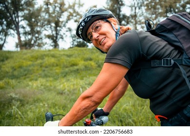 Senior Woman Cyclist In Black Sportswear And Helmet Riding Bicycle Outdoors In Green Nature. Copy Space