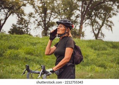 Senior Woman Cyclist Using Mobile Phone In The Green Wild Nature. Elderly Female In Black Sports Clothes And Cycling Helmet Talking On A Phone Outdoors. Copy Space