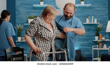 Senior Woman With Disability Receiving Help From Man Nurse Living In Nursing Home, Getting Dressed With Jacket. Healthcare Specialist Giving Assistance To Retired Patient With Walk Frame