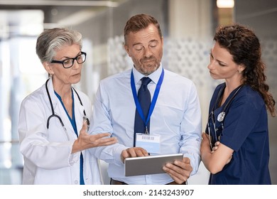 Senior woman doctor having a discussion in hospital hallway with medical staff. Doctor discussing patient case status with his team. Pharmaceutical representative showing medical report on tablet.