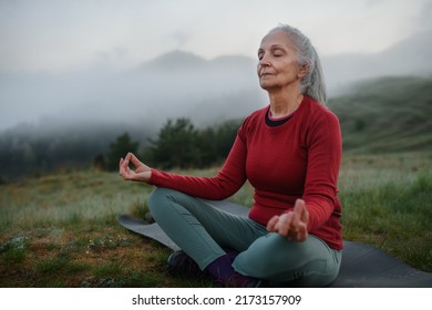 Senior woman doing breathing exercise in nature on early morning with fog and mountains in background. - Powered by Shutterstock