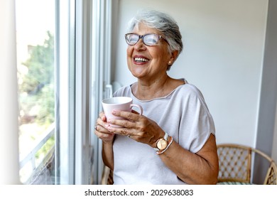 Senior woman drinking a beverage at home. Charming elderly woman posing by window and smiling while drinking coffee from a pink cup. Old woman is staying at home during the COVID-19 outbreak. - Powered by Shutterstock