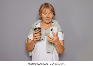 Senior woman drinking coffee chatting on her mobile addicted to social media reading news searching online browsing web page doing shopping in web store standing isolated over background - Powered by Shutterstock