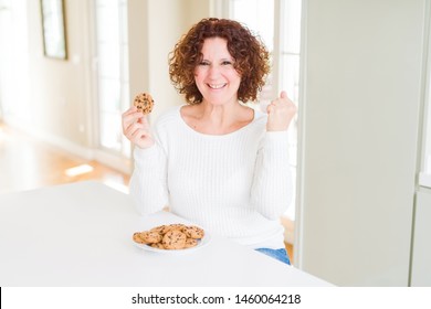 Senior woman eating chocolate chips cookies at home screaming proud and celebrating victory and success very excited, cheering emotion