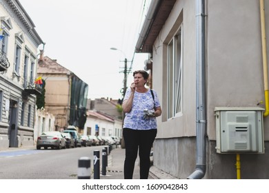 Senior Woman Eating Chocolate While Walking On The Sidewalk - One Old Person Biting And Enjoying A Cocoa Bar Outside