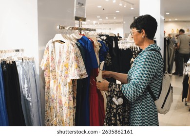 Senior woman enjoying leisure time while browsing through clothing racks in a fashion retail store, checking price tags and fabrics as she selects garments for purchase