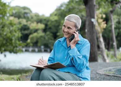 Senior Woman Enjoying Outdoor Lifestyle While Engaging in a Phone Call and Taking Notes in a Serene Park Setting - Powered by Shutterstock