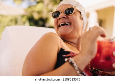 Senior woman enjoying a tiki cocktail while vacationing in the summer. Carefree elderly woman laughing cheerfully while relaxing on a lounger. Happy mature woman enjoying herself after retirement. - Powered by Shutterstock