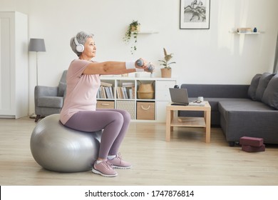 Senior woman exercising with dumbbells while sitting on fitness ball and listening to music in the room at home - Powered by Shutterstock