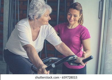 Senior woman exercising on stationary bikes in fitness class. Woman workout in gym. Senior with personal trainer. - Powered by Shutterstock