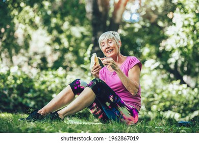 Senior Woman Exercising In Park While Listening To Music. Mature Woman Eat Banana Resting After Exercise.