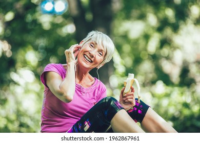 Senior Woman Exercising In Park While Listening To Music. Mature Woman Eat Banana Resting After Exercise.