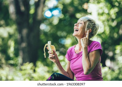 Senior Woman Exercising In Park While Listening To Music. Mature Woman Eat Banana Resting After Exercise.