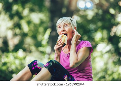 Senior Woman Exercising In Park While Listening To Music. Mature Woman Eat Banana Resting After Exercise.