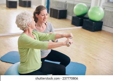 Senior Woman Exercising With Weights In The Gym Assisted By A Young Female Trainer. Old Woman Lifting Dumbbells With Help From Personal Trainer At Rehab.