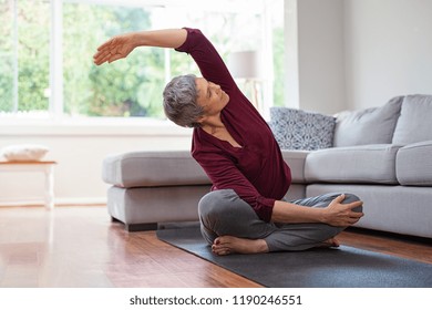 Senior Woman Exercising While Sitting In Lotus Position. Active Mature Woman Doing Stretching Exercise In Living Room At Home. Fit Lady Stretching Arms And Back While Sitting On Yoga Mat.