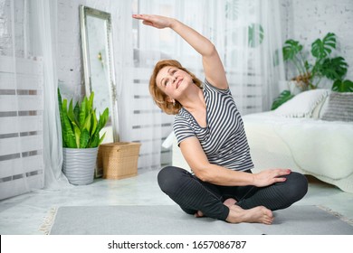 Senior Woman Exercising While Sitting In Lotus Position. Active Mature Woman Doing Stretching Exercise In Living Room At Home.