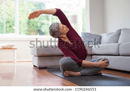 Senior woman exercising while sitting in lotus position. Active mature woman doing stretching exercise in living room at home. Fit lady stretching arms and back while sitting on yoga mat.