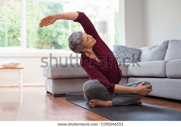 Senior woman exercising while sitting in lotus\
position. Active mature woman doing stretching exercise in living\
room at home. Fit lady stretching arms and back while sitting on\
yoga mat.