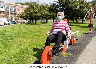 Senior Woman With Face Mask Doing Exercise On Static Bicycle Machine At Park On Sunny Day. Elder Lady Training During Pandemic To Keep Fit And Healthy Outdoors