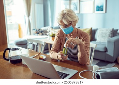 Senior woman in face mask holding medication using laptop at home - Powered by Shutterstock