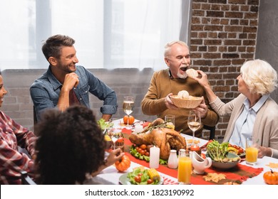 Senior Woman Feeding Man With Bun Near Multicultural Family During Thanksgiving Dinner