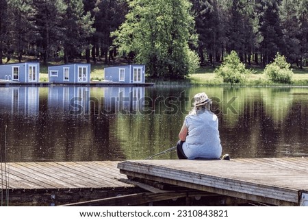 Senior woman with a fishing rod sitting on a wooden pier by a pond with reflections on a sunny day in summer