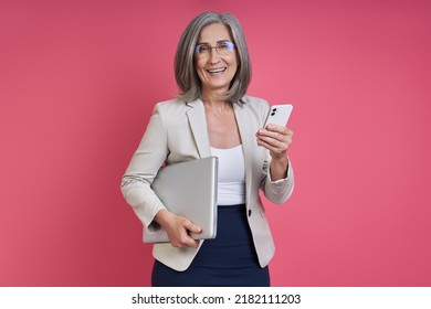 Senior woman in formalwear carrying laptop and smart phone while standing against pink background - Shutterstock ID 2182111203