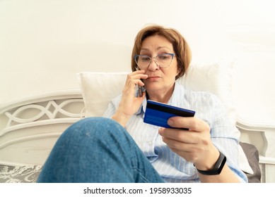 Senior woman frustrated talking on mobile phone indoors and looking on bank card in her hand