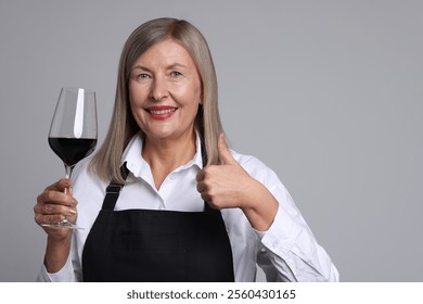 Senior woman with glass of wine showing thumbs up on grey background - Powered by Shutterstock