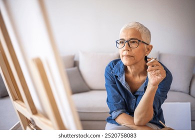 A senior woman with glasses deeply focused on painting in a cozy living room. She exudes creativity and concentration, showcasing hobbies and artistic expression in a relaxed environment. - Powered by Shutterstock