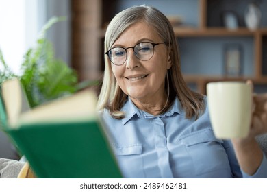 Senior woman with glasses reading book and drinking coffee in living room. Relaxed and content. Concept of leisure, happiness, and retired life. Comfortable cozy home environment with natural light - Powered by Shutterstock