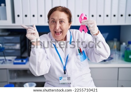 Senior woman with glasses working at scientist laboratory holding pink ribbon smiling happy pointing with hand and finger to the side 