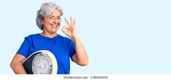 Senior Woman With Gray Hair Holding Weight Machine To Balance Weight Loss Doing Ok Sign With Fingers, Smiling Friendly Gesturing Excellent Symbol 