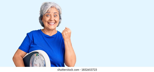 Senior Woman With Gray Hair Holding Weight Machine To Balance Weight Loss Screaming Proud, Celebrating Victory And Success Very Excited With Raised Arms 