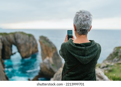 Senior woman with gray hair using her smartphone to take a landscape photo while hiking along a coastal path. - Powered by Shutterstock