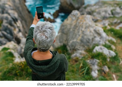 Senior woman with gray hair using her smartphone to take a landscape photo while hiking along a coastal path. - Powered by Shutterstock