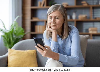 Senior woman with gray hair using smartphone, sitting on sofa, looking thoughtful. She appears relaxed and engaged with technology, possibly checking messages or social media on phone. - Powered by Shutterstock