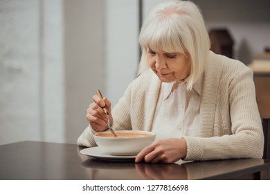 Senior Woman With Grey Hair Sitting At Table And Eating Cream Soup At Home 