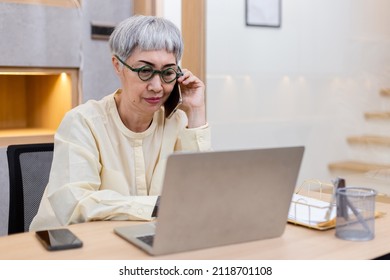 Senior Woman Grey Hair Speaking On Mobile Phone Using Laptop At Office Table. Elderly Businesswoman Working On Laptop Computer.