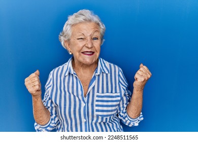 Senior woman with grey hair standing over blue background very happy and excited doing winner gesture with arms raised, smiling and screaming for success. celebration concept.  - Shutterstock ID 2248511565