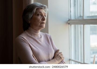 A senior woman with grey hair is standing by a window. She is wearing a pink sweater and has her arms crossed. The woman looks contemplative as she gazes out of the window. - Powered by Shutterstock